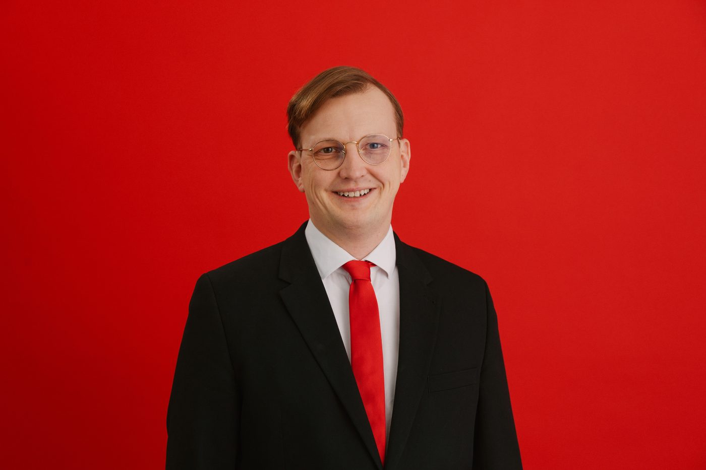 Portraitfoto von Alexander Mittag, dem Oberbürgermeisterkandidaten der SPD für Delmenhorst bei der niedersächsischen Kommunalwahl im September 2026. Er trägt einen schwarzen Anzug, ein weißes Hemd und eine Krawatte in SPD-rot. Der Hintergrund ist ebenfalls im gleichen Feuerrot. Die Haare sind zu einem Seitenscheitel gekämmt und er lächelt freundlich in die Kamera.