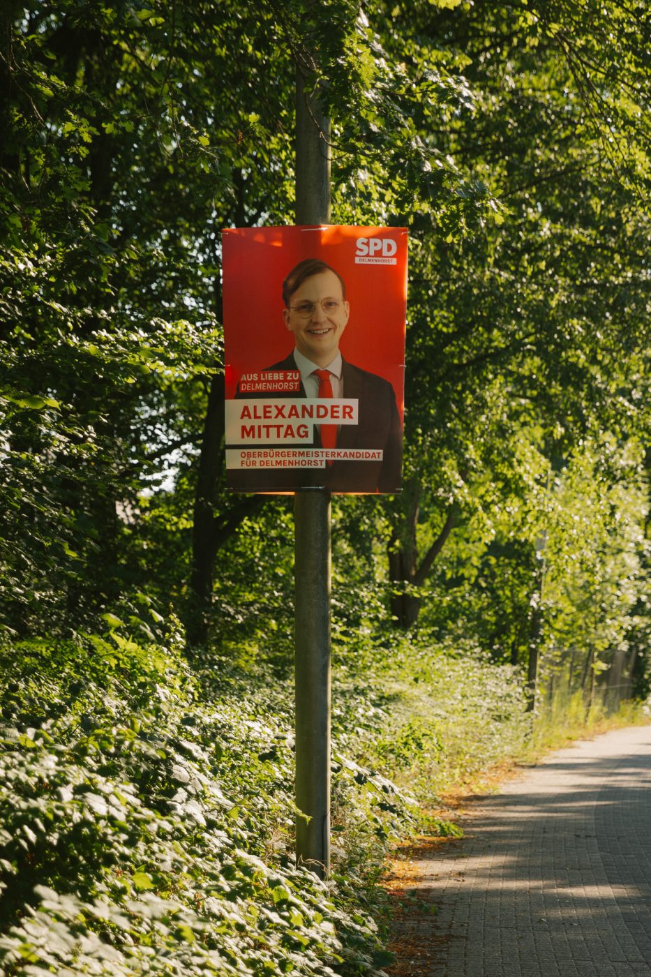 Ein Wahlplakat für Alexander Mittag von der SPD als Oberbürgermeister-Kandidat. Das Plakat hängt an einem Laternenmast an einer Straße, im Hintergrund sind Büsche und Bäume zu sehen.