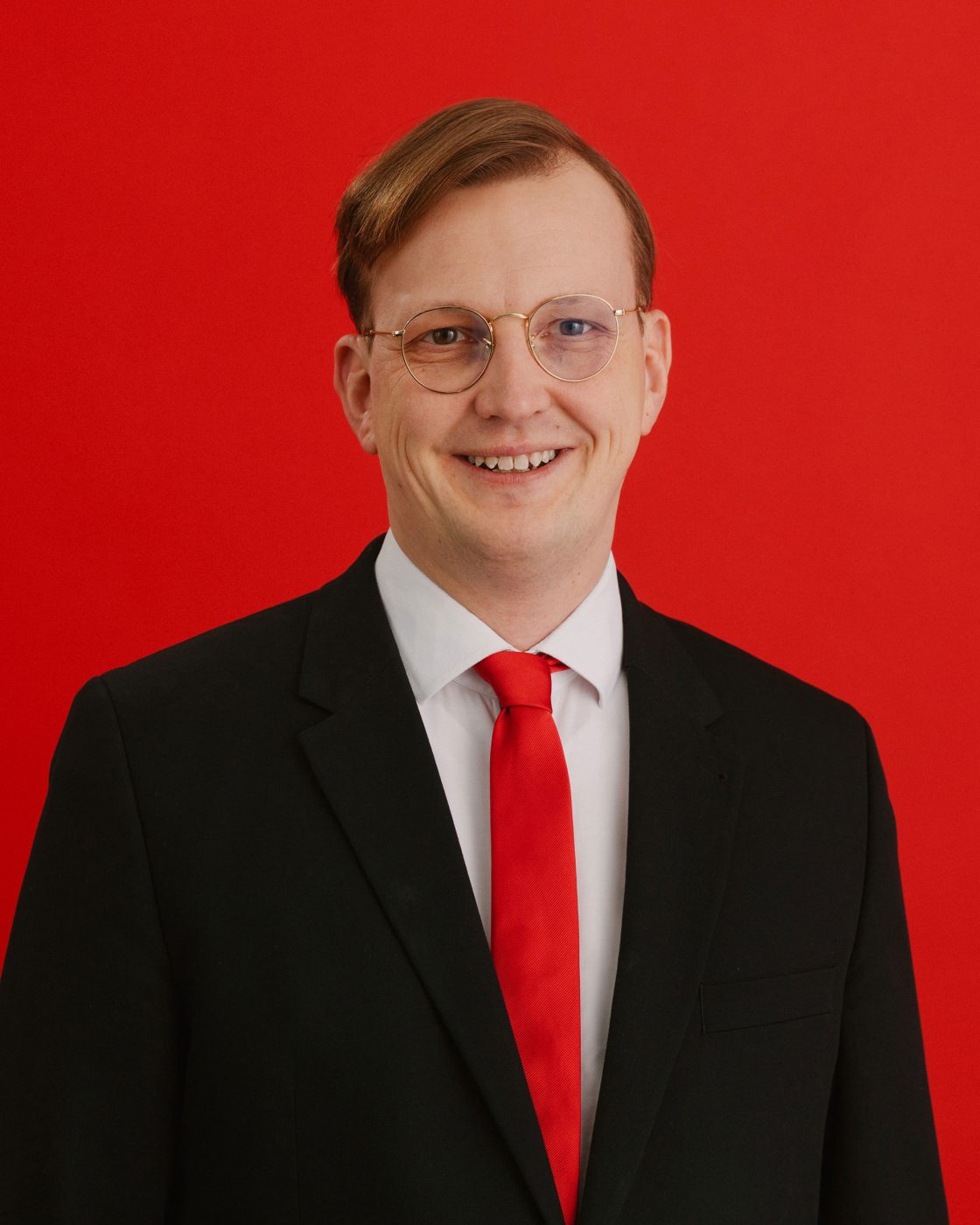 Portraitfoto von Alexander Mittag, dem Oberb&uuml;rgermeisterkandidaten der SPD f&uuml;r Delmenhorst bei der nieders&auml;chsischen Kommunalwahl im September 2026. Er tr&auml;gt einen schwarzen Anzug, ein wei&szlig;es Hemd und eine Krawatte in SPD-rot. Der Hintergrund ist ebenfalls im gleichen Feuerrot. Die Haare sind zu einem Seitenscheitel gek&auml;mmt und er l&auml;chelt freundlich in die Kamera.